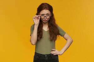 Cute, flirty woman with long ginger hair. Wearing green t-shirt and glasses. People and emotion concept. Touching her eyewear and lifts eyebrow. Watching at the camera, isolated over orange background photo
