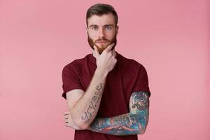 A confident man with a beard and with tattooed hand, holds his hand on chin, thinks about something, build a plan, ponders a cool idea. Isolated over oink background. photo