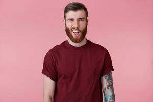 Portrait of young bearded male with tattooed hand makes a frown, screwing eye, isolated over pink background. photo