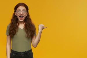 Excited, amazed woman with long ginger hair. Wearing green t-shirt and glasses. Watching at the camera and pointing with thumb to the right at copy space, isolated over orange background photo