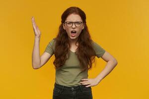 Portrait of unhappy, pissed lady with long ginger hair. Wearing green t-shirt and eyewear. People and emotion concept. Raises hand and arguing. Watching at the camera, isolated over orange background photo