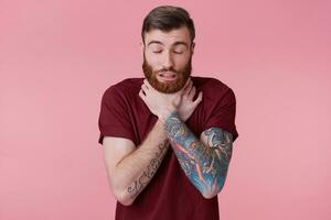 Close up of young bearded inked man choking himself, experiencing severe sore throat, isolated over pinkground. photo