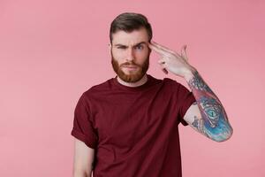 Photo of young bearded inked man isolated over pink background, shooting at his head with hand gun, showing suicide gesture.