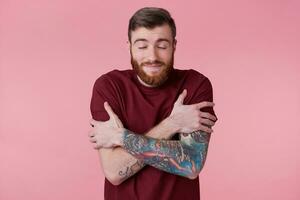 Photo of happy young bearded man with inked hand, with closed eyes, hugs himself, think about something cute. Isolated over pinkbackground.