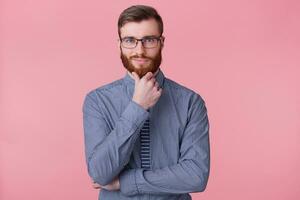 A confident young bearded man in glasses, holds his hand on chin, thinks about something, build a plan, ponders a cool idea. Isolated over oink background. photo