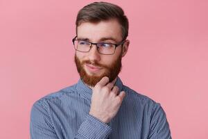 Close up of young bearded businessman with serious face expression thinking about question, thoughtfully touching his chin, looking into the distance isolated over pink background. photo