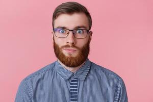 Close up portrait of funny ginger guy in glasses and striped shirt looking at camera, isolated on pink background photo