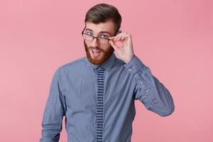 Portrait of a young handsome bearded man who tells a funny story, looks through his glasses isolated over pink background. photo