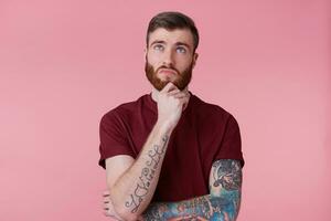 Close up photo of thinking young bearded male with tattooed hand, holding fist on chin, looking aside isolated over pink background. Waiting for inspiration.