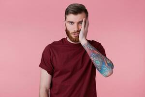 Portrait of sad bored young man with tattooed hand, propping up his head, isolated over pink background. photo