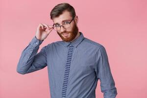 Close up of young handsome bearded man posing at camera isolated over pink background and smiling, holding his glasses with hand and looking through them. photo