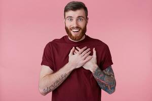 Portrait of happy amazed young bearded man with tattooed hand, putting palms to the heart, smiles broadly and looking at the camera, isolated over pink background. photo