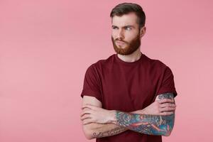 Portrait of displeased beautiful young man with ginger beard and tattooed hand, holding hands crossed, frown and looking away isolated over pink background. People and emotion concept. photo