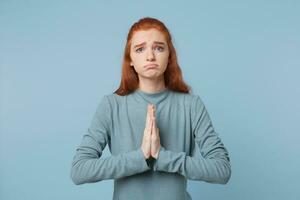 Red-haired girl prays asking for something. A teenager with a pitifully sad expression on her face folded her arms in front of herself showing a begging pleading gesture isolated over blue background photo