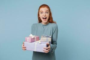 Happy birthday girl ready to scream with joy and happiness delight. Red-haired girl holding boxes with gifts in her hands, stands sideways with wide open eyes and mouth, isolated on a blue background. photo