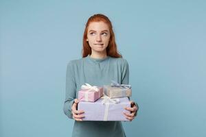 Cute thoughtful young red-haired girl stands holding boxes with gifts, looks away, bites her lip, thinks about something, ponders, remembers something, isolated on a blue background. photo