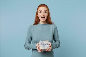 Red-haired girl overwhelmed with positive emotions, insanely happy, joyful wide-open eyes and mouth, holding a wrapped gift tied with a blue ribbon isolated on a blue background photo