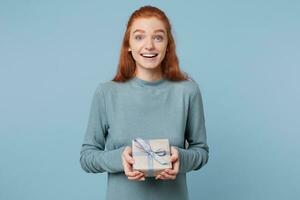 A young red-haired girl received a wrapped gift tied with a blue ribbon holding it in her hands smiling glows from happiness isolated on a blue background. photo