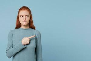Red-haired girl looks to the side with suspicion cautiously biting her lip shows with her index finger to the empty space for copy promotion advertisement, isolated on a blue background. photo