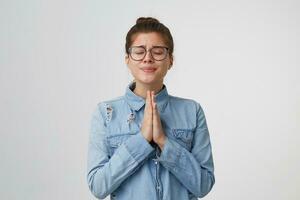 un bonito joven niña en lentes soportes con ojos cerrado, manos doblada en frente de su, Orando gesto, preguntando para alguna cosa importante, falto, esperando para alguna cosa aislado en blanco antecedentes foto