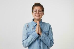 Portrait of young girl in glasses stands with eyes closed, hands folded in front of her, praying gesture, asking for something important gritting teeth hard, wanting, hoping for something photo