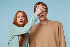 A guy and a girl isolated on a blue background. The girl looks into the camera. Scaredly shocked, she closes the guy's eyes with her palm, as if she sees something indecent, immoral and propriety. photo