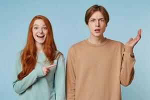 Couple,girl with long red hair laughs shows with her index finger at the guy who stands with a bewildered, confused, puzzled expression,hand raised up,isolated on blue background photo