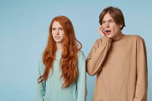 A guy and a red-haired girl are standing sideways looking distance look at something unpleasant with disgust, horrified, the guy wrinkles his face and holds his hand to his cheek photo