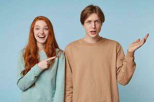 Couple,girl with long red hair laughs shows with her index finger at the guy who is standing with an uncomprehending frown expression outraged,something says,hand raised up,isolated on blue background photo