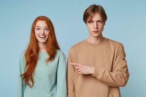 Close up couple, girl smiles joyful happy with wide open eyes and a smile,long red hair, the guy is not in the spirit displeased points his index finger to his girlfriend,isolated on a blue background photo