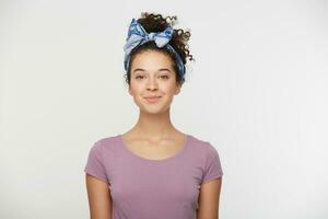 Happy female with positive expression, dressed in casual t shirt and stylish headband, poses against white background, looking at the camera with a playful smile. People and emotions photo