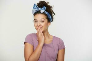 Positive young female closed mouth with hand, looks playfully, dressed in casual t shirt and headband, flirt. Isolated over white background photo