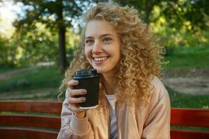 al aire libre foto de joven hermosa niña rubia se sienta en un parque banco, Bebiendo café, mira en un compañero, lejos, Hablando con alguien, sonriente, siente feliz, en un bueno compañía, tiene un bueno tiempo, soñando