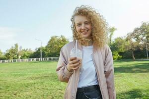 hermosa joven niña rubia con Rizado pelo poses en el parque, sonriente, viento olas su cabello, bebidas un cóctel, un zalamero, vestido por casualidad en un rosado chaqueta terminado un blanco camiseta, mira cámara foto