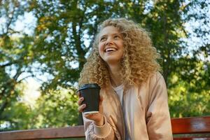 un joven hermosa niña rubia se sienta en un parque banco, Bebiendo café, mira en un compañero, Hablando con alguien, reír, siente feliz, en un bueno compañía, tiene un bueno hora foto