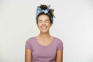 Laughing from happiness female, positive expression, dressed in casual t shirt and stylish headband, poses against white background, closed eyes. People and emotions. Joyful long-awaited moment photo