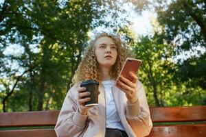 un joven hermosa niña rubia se sienta en un parque banco, Bebiendo café, mira en un compañero, Hablando con alguien, sostiene el teléfono en mano, reflexiona el leer mensaje, ver desde abajo, parque antecedentes. foto