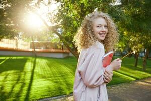Beautiful curly hair young girl passes by with a red notebook and a pencil in hands, headphones in her ears with favorite music, looks into the camera and smiles, park background. photo