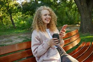 Outdoor photo of young beautiful girl blonde sits on a park bench, drinking coffee, holds her phone in hand, ponders the read message, smiling, feels happy, dressed pink jacket, side view