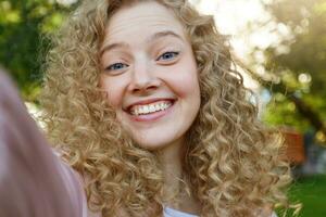 Young beautiful funny cool girl blonde with curly hair, widely smiling and looking into the camera, making selfie, nature background photo
