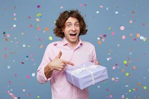 Exited happy birthday guy got a surprise party, posing for picture, holding box with present and pointing on it with index finger, confetti falling down. People, joy, fun and happiness concept. photo