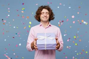 People, joy, fun and happiness concept. Relaxed happy birthday guy with eyes closed waiting for a surprise, posing for picture, holding box with present and with confetti falling down photo