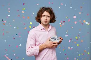 Guy posing with a gift box looks as if he is afraid that someone will take it away, does not want to part with it, holds present tightly in his hands, against the background of falling confetti. photo