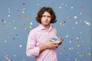 Guy posing with a gift box looks as if he is afraid that someone will take it away, does not want to part with it, holds present tightly in his hands, against the background of falling confetti. photo