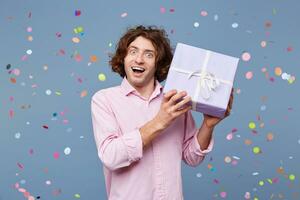 Birthday man accepts congratulations, received a gift in a box, shows it, poses joyfully inspired, smiles, eyes and mouth wide open, confetti falling down. photo