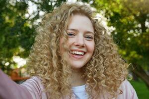 Selfie photo of young beautiful lovely girl blonde with curly hair, widely smiling, looks happy, nature background