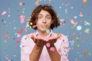 Close up of happy male blowing confetti off his hands. Party man gladly standing around falling down confetti, over blue background photo