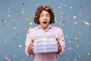 Cheerful happy young male man with a decorative gift isolated on blue background. Man is receiving gift box. He is standing and looking at camera with surprise. photo