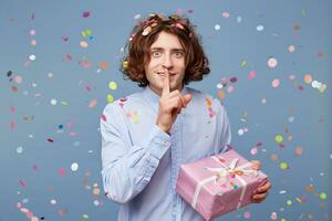 Nice cute young man having secret, holding finger on lips and showing silence sign, standing on blue background.Emotional male with long hair ask to keep silence, holding a gift box photo