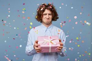 Portrait of a guy holding a present box with a frustrated expression, pursed his lips, dissatisfied, upset, offended, looking into the camera standing against the blue background of falling confetti. photo